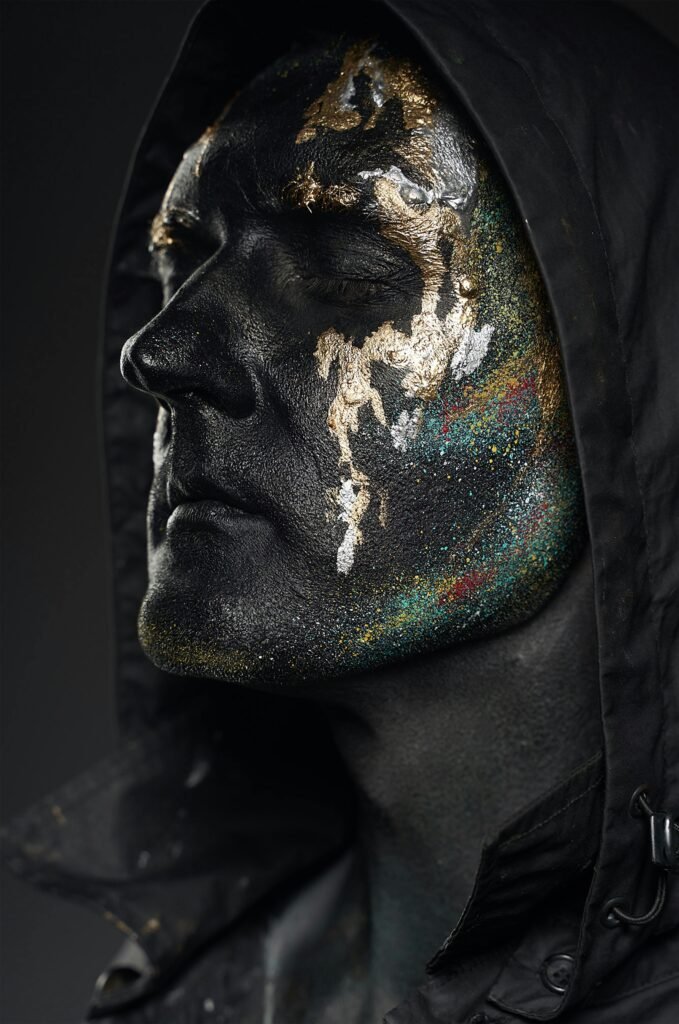 Close-up artistic portrait of an adult with black and gold face paint in a moody setting.